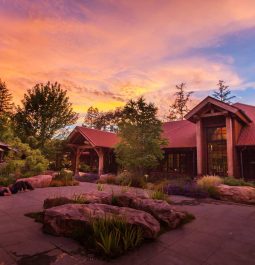 Yellow and purple sunset behind an expansive two story lodge surrounded by trees. A woman lies on her back in the stone terrace in front.