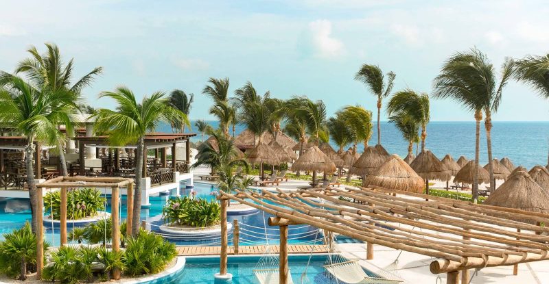 A luxurious tropical resort with a serene blue pool, surrounded by lush palm trees, thatched-roof cabanas, and hammocks. In the background, there's a calming view of the ocean under a clear blue sky.